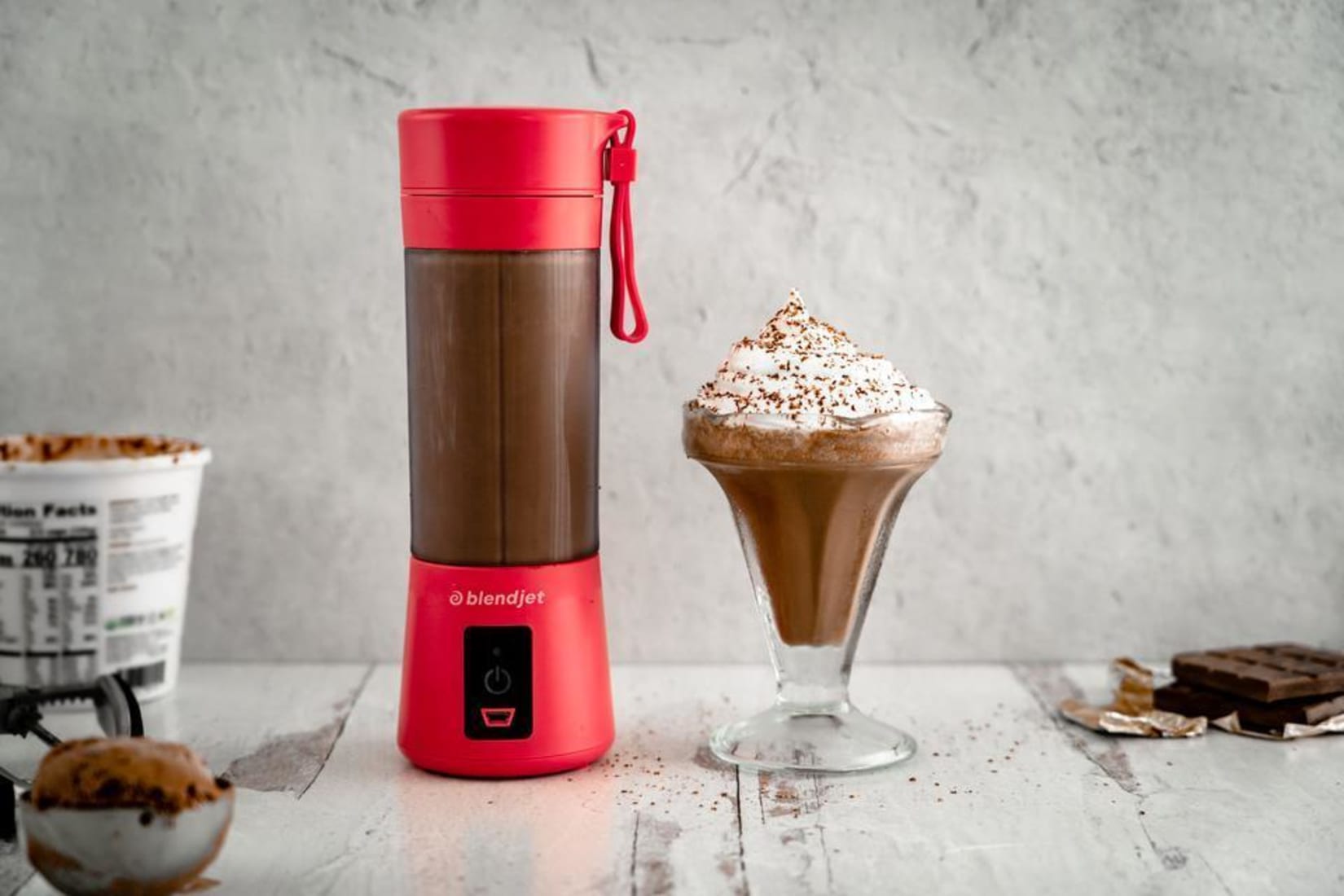 Red BlendJet on a counter next to a chocolate shake and surrounded by chocolate and cocoa powder